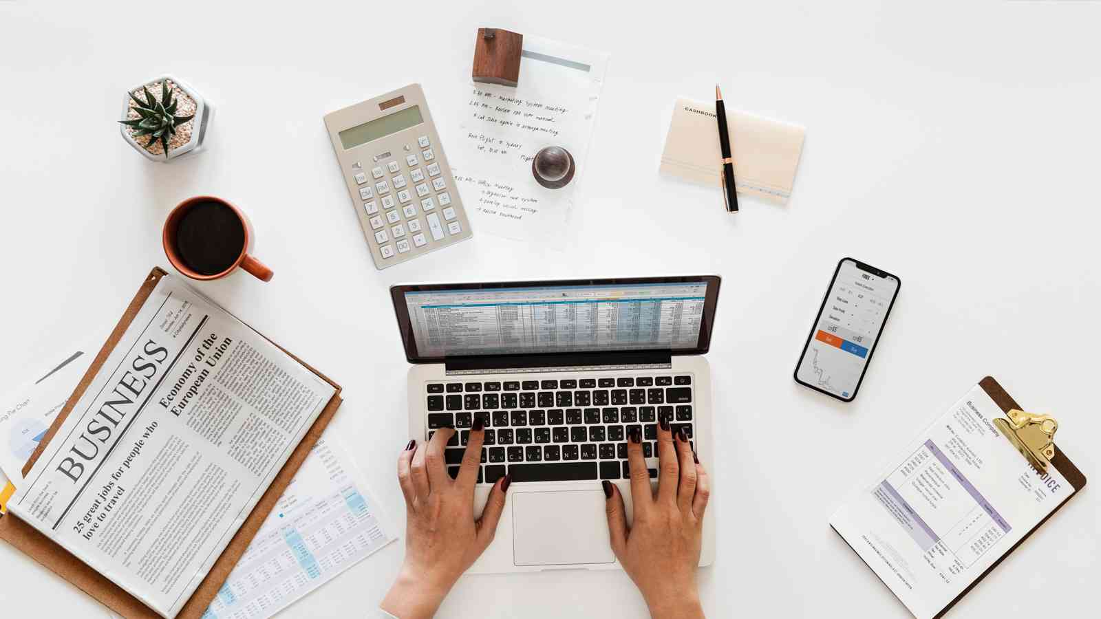 Top-down view of a person typing on a laptop, surrounded by business documents, a calculator, a smartphone displaying a graph, a newspaper, a cup of coffee, and office supplies on a white desk.
