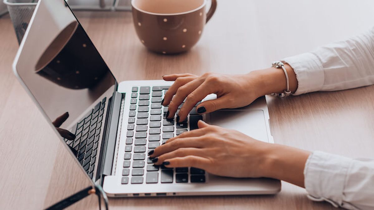 Close-up of a woman's hands typing on a laptop keyboard, with a polka-dotted coffee mug in the background on a wooden desk.