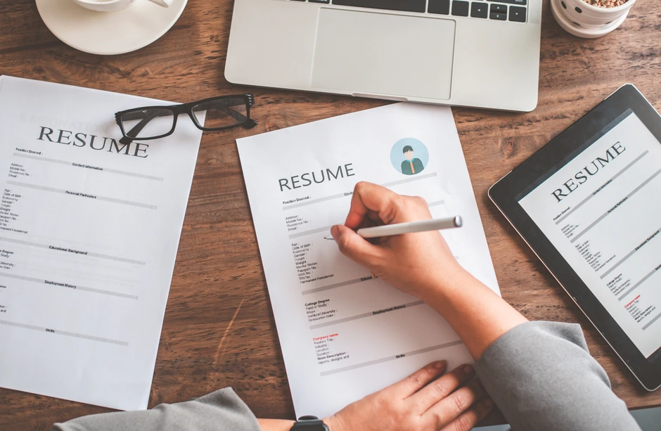 Common Resume Mistakes to Avoid A professional reviewing and editing a printed resume at a wooden desk with a laptop, tablet, and eyeglasses.