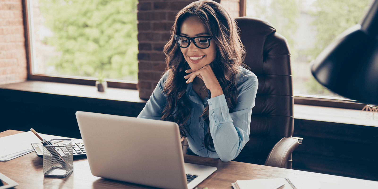 WFH3 A professional woman with glasses and long wavy hair is smiling while working on her laptop in a well-lit home office. She is wearing a light blue shirt and sitting in a leather chair, surrounded by office essentials like papers, a pen holder, and a desk lamp
