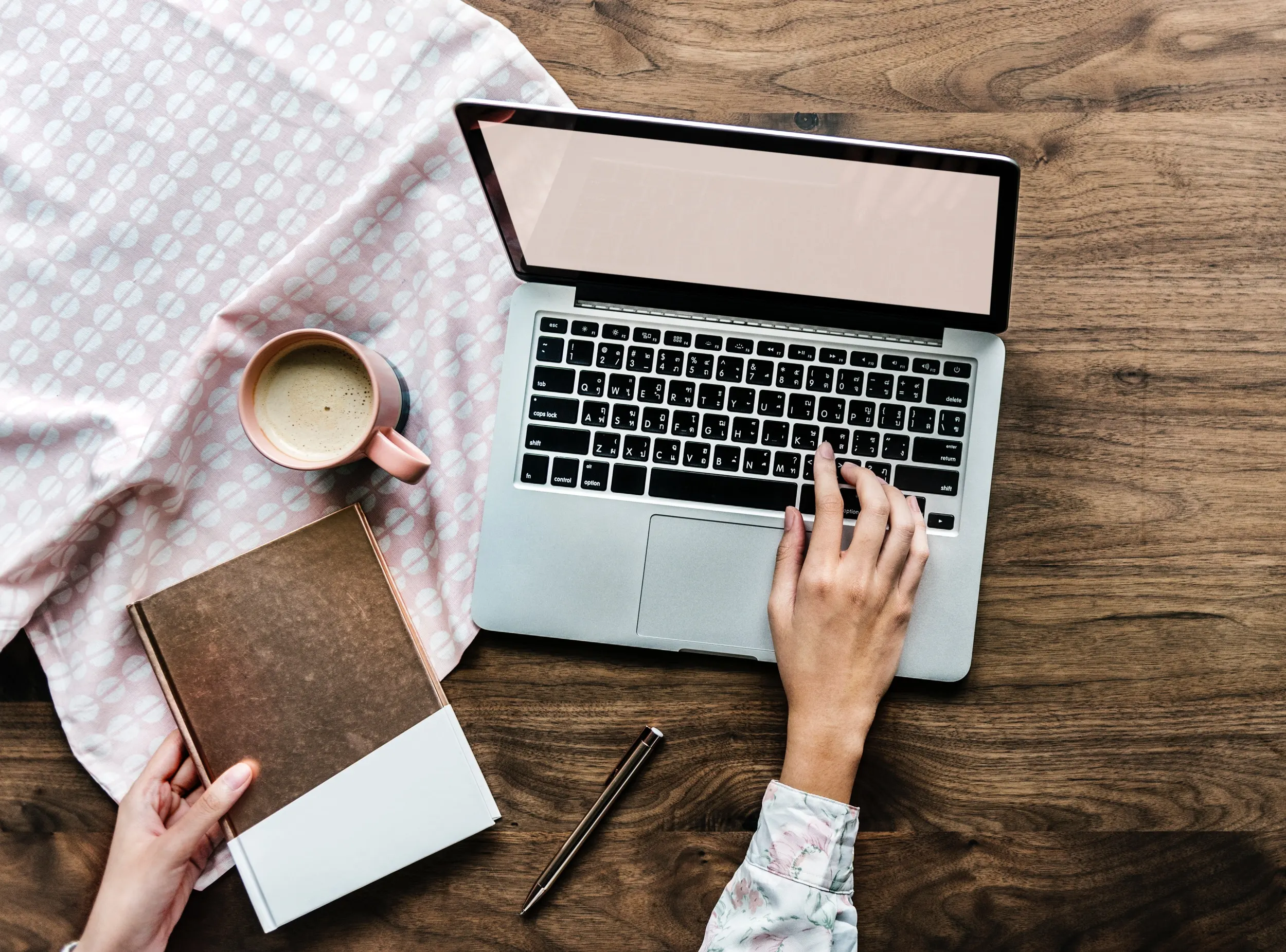 WFH4 A top-down view of a woman working on a laptop at a wooden desk, with one hand on the keyboard and the other holding a book. A cup of coffee, a pen, and a patterned cloth are also on the desk.