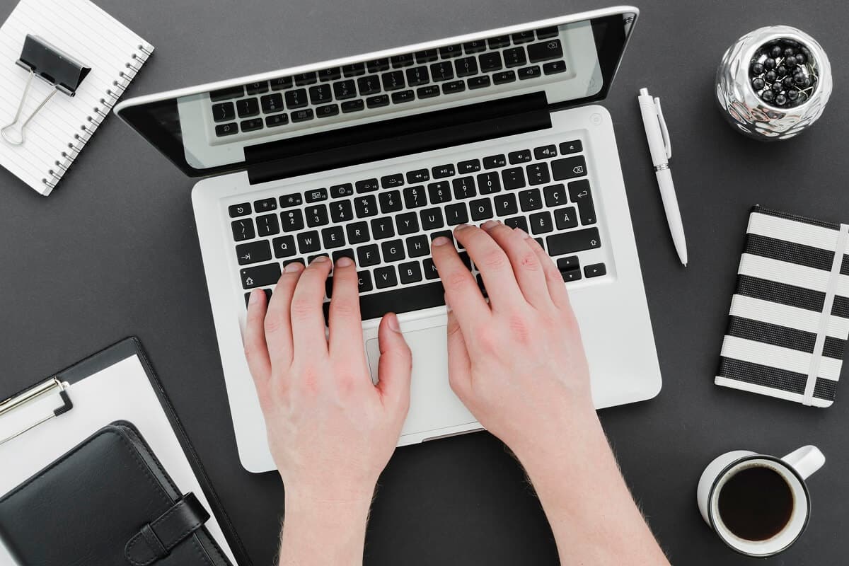 Top view of a person typing on a laptop keyboard, surrounded by office supplies, a notebook, and a cup of coffee on a black desk.